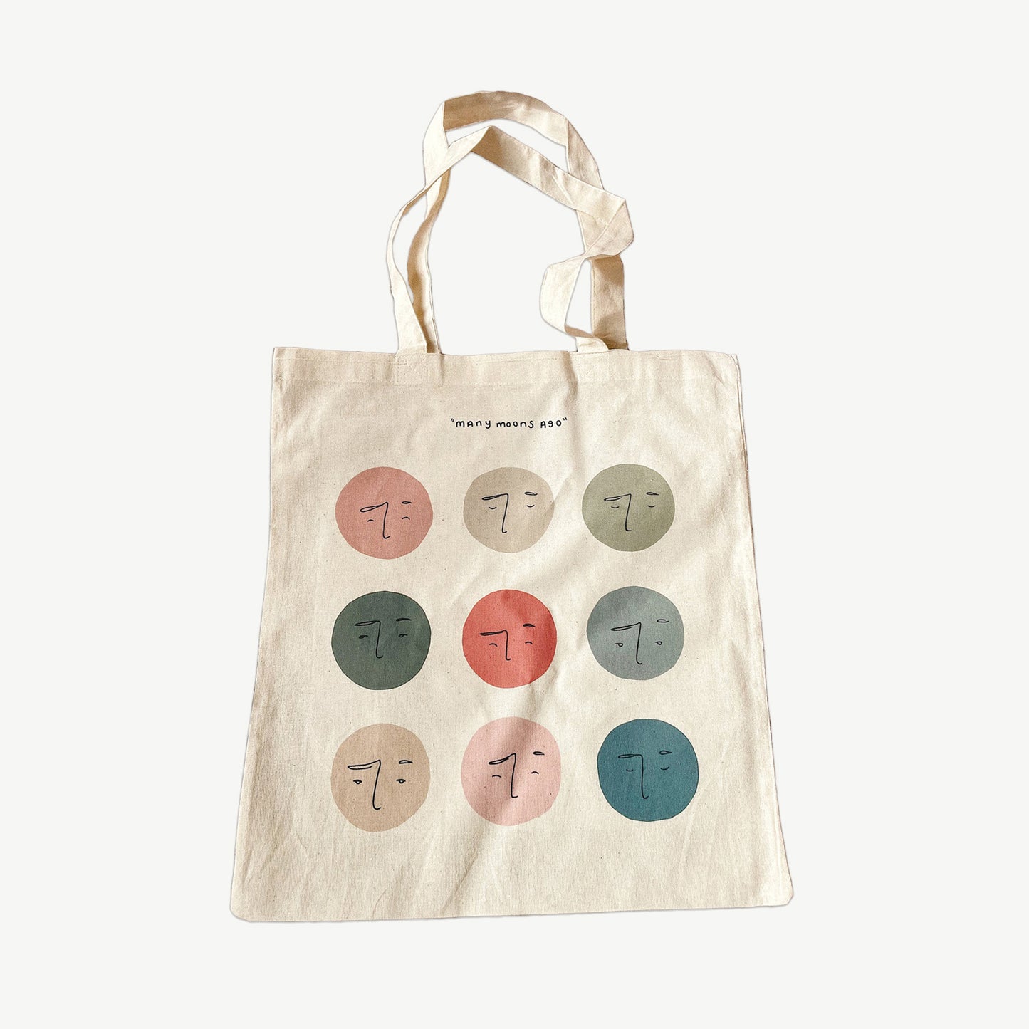 A Cotton Tote bag with nine hand drawn moons in multiple colours, with the words 'many moons ago' written across the tote in black writing. Recycled cotton tote bag 